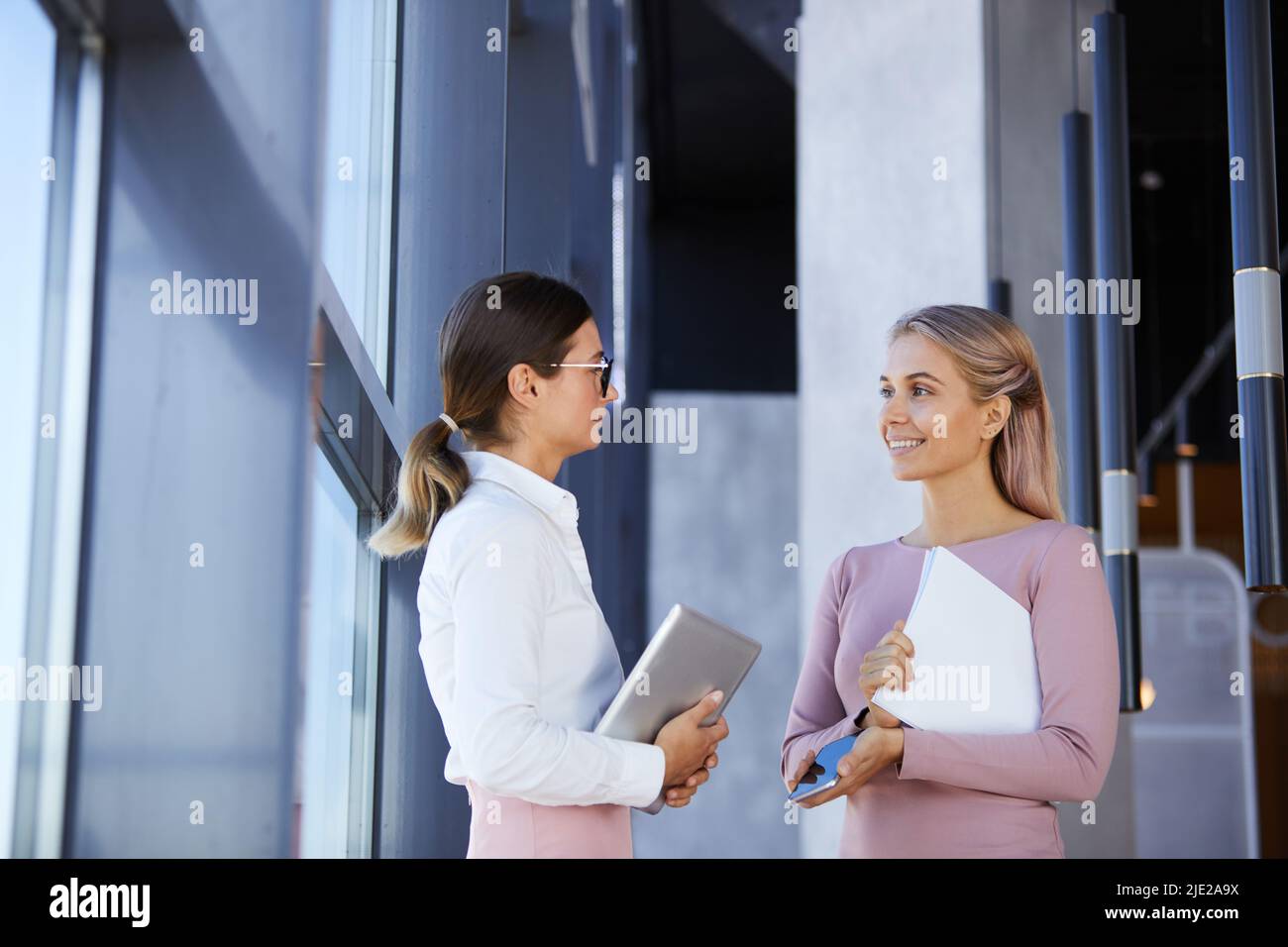 Des collègues féminins positifs et attrayants dans des tenues élégantes debout à la fenêtre dans le couloir et parlant de travail Banque D'Images