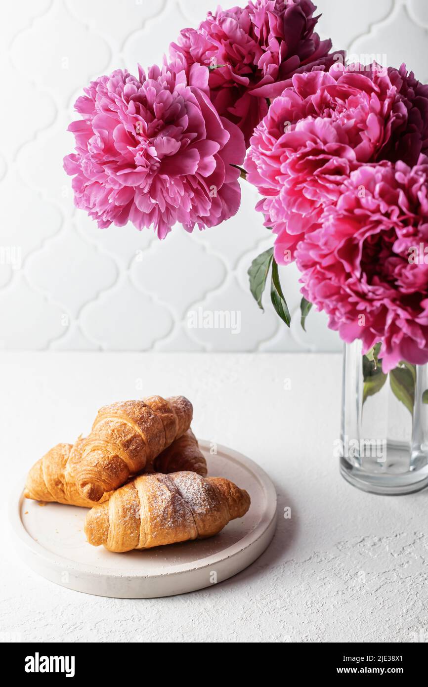 Croissants dans une assiette et bouquet de pivoines roses sur une table blanche. Esthétique encore la vie. Orientation verticale Banque D'Images