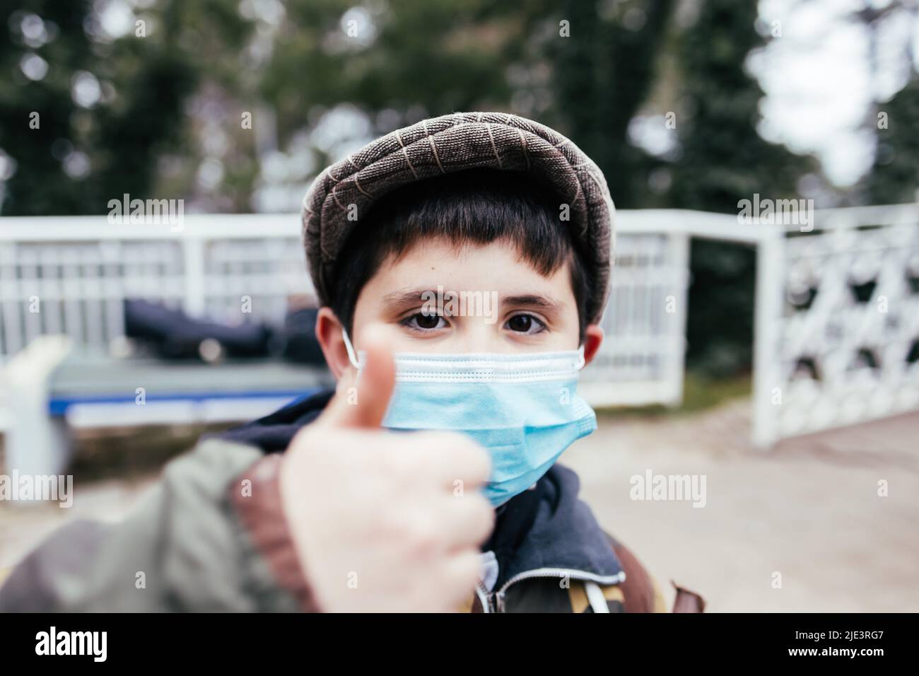 Petit enfant avec un béret et un masque facial sur faire le signal de pouce vers le haut, au parc. Banque D'Images