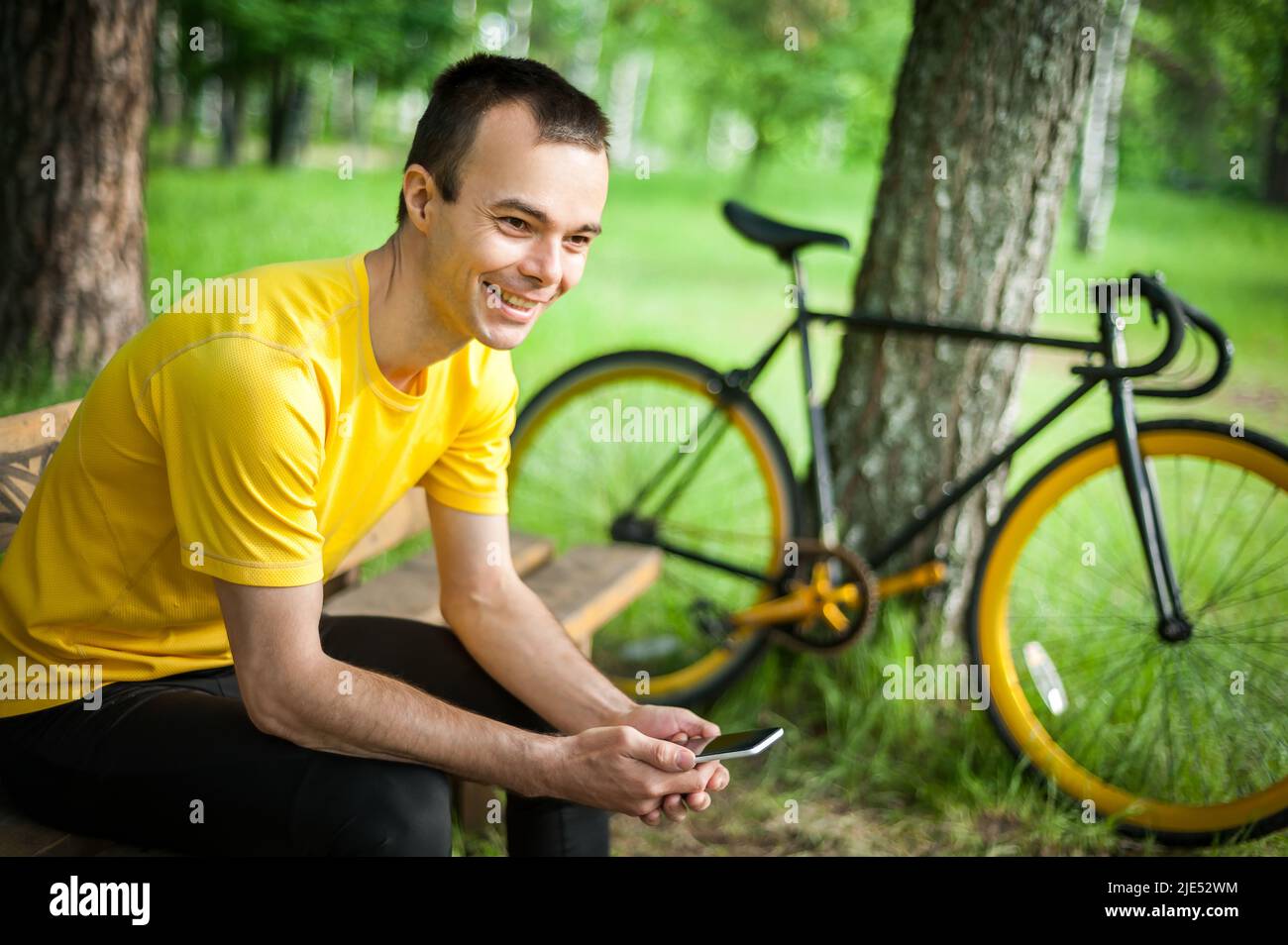 Un jeune homme assis sur un banc dans un parc public communique par communication mobile. En arrière-plan avec son vélo. Parmi les arbres et la végétation. Banque D'Images