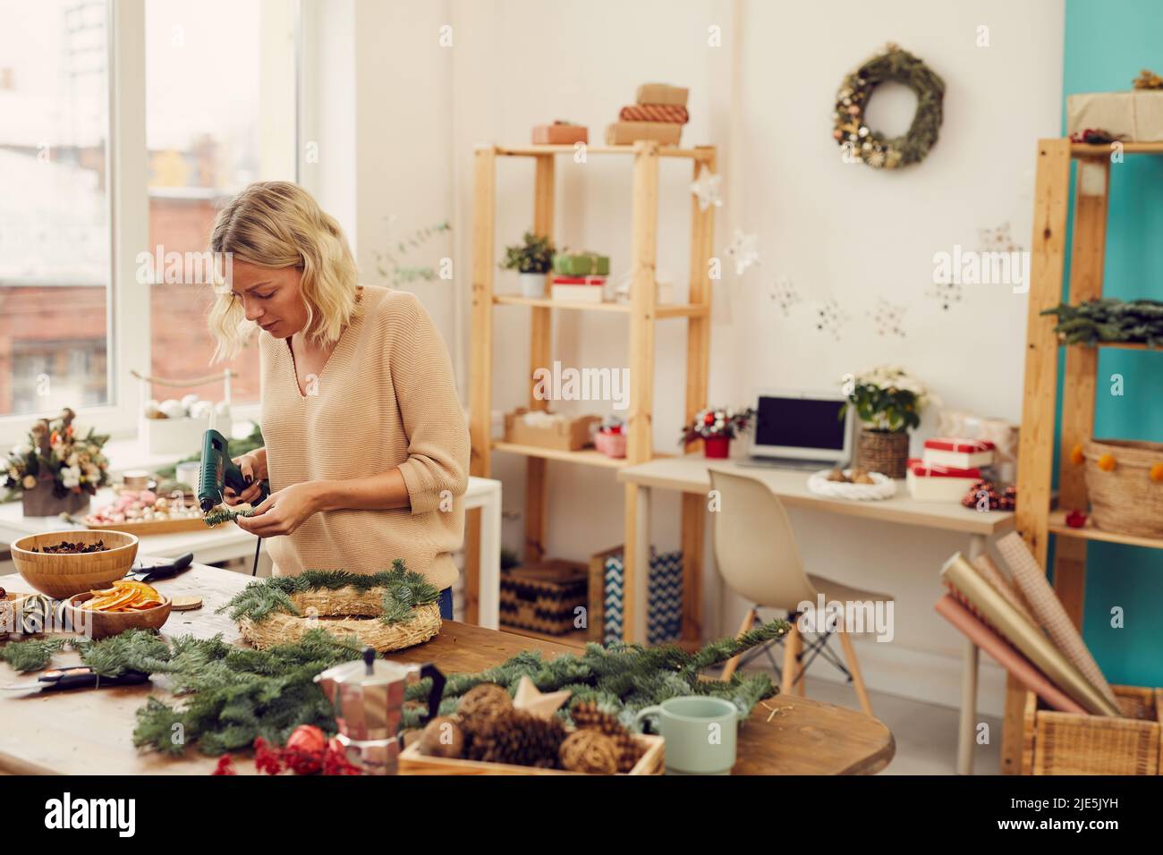 Jeune artisan concentré attrayant avec cheveux blonds debout à la table et à l'aide d'un pistolet à colle chaude tout en créant une couronne de Noël en studio Banque D'Images