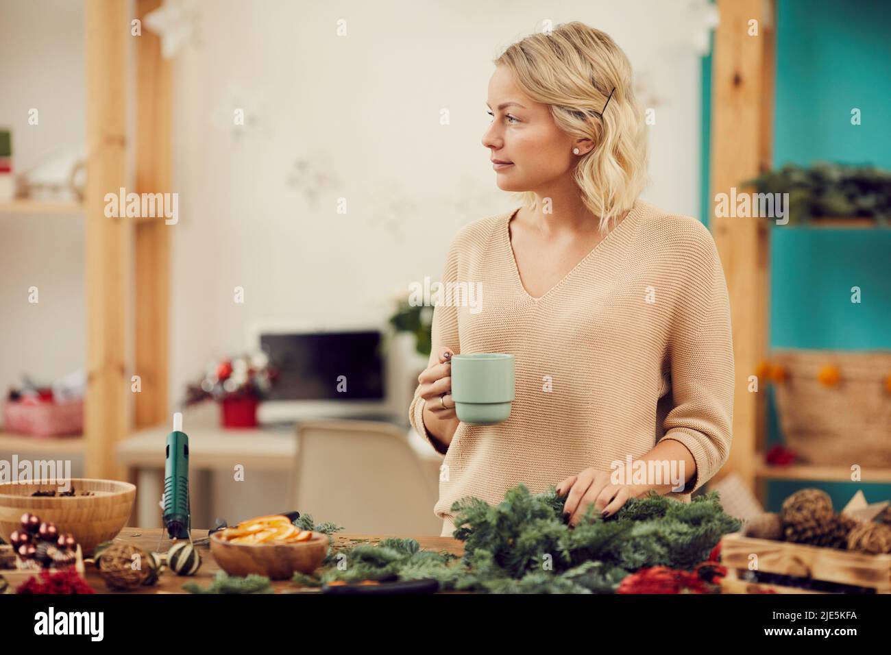 Jolie jeune femme pensive aux cheveux blonds ondulés debout sur le bureau avec des décorations de Noël et regardant loin avec soin tout en buvant du café à l'intérieur Banque D'Images