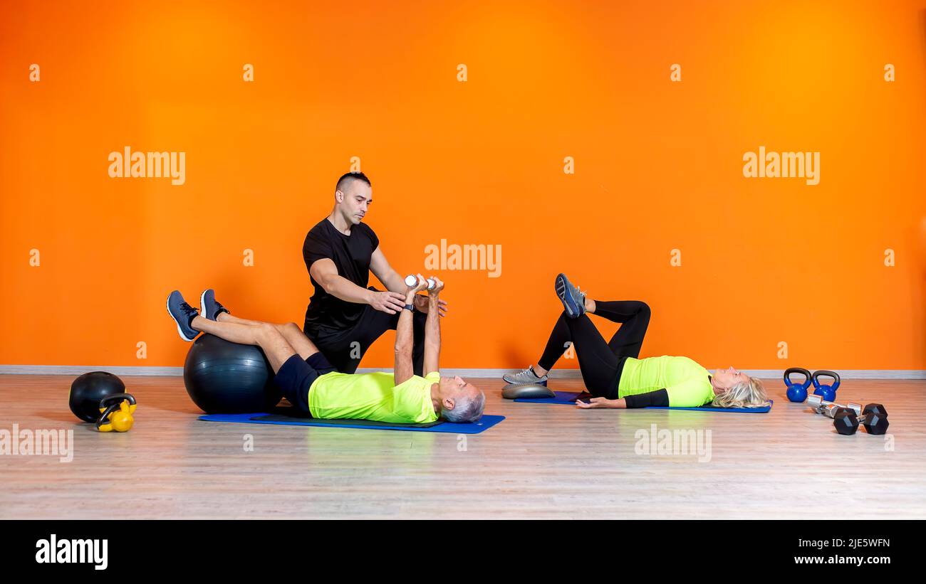 senior fit couple faire l'exercice de musculation exercice d'entraînement se posant à la salle de gym isolé sur fond orange avec entraîneur personnel. personnes matures fitness Banque D'Images