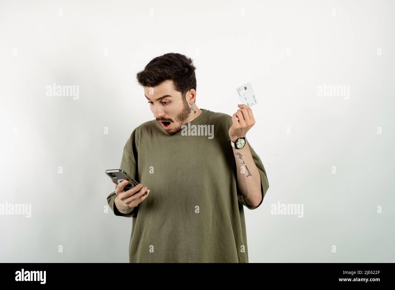 Beau homme caucasien portant un t-shirt kaki posant isolé sur fond blanc tenant smartphone et carte de crédit choqué et étonné Banque D'Images