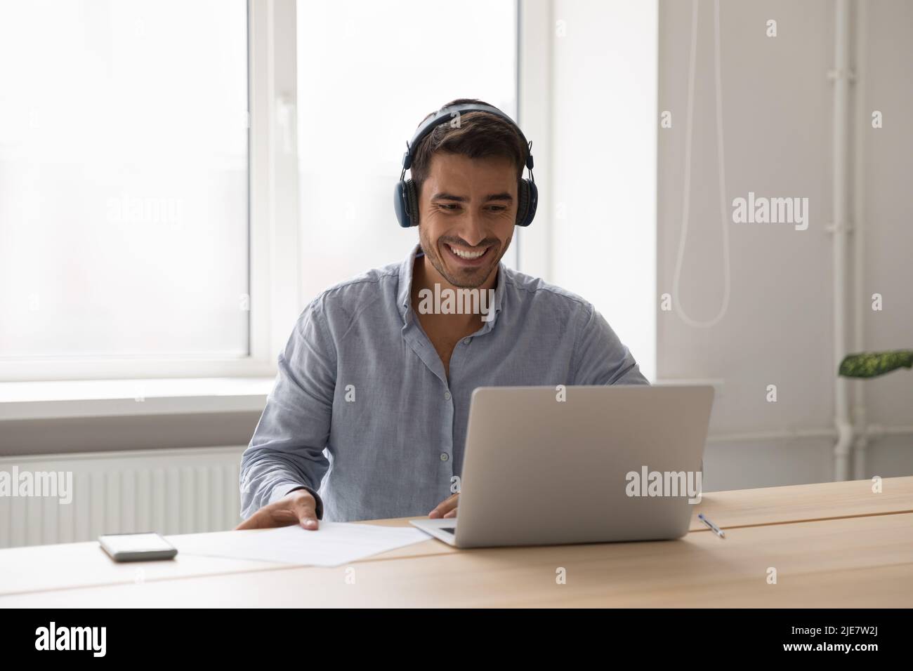 Un homme d'affaires souriant porte un casque, assis au bureau avec un ordinateur portable Banque D'Images