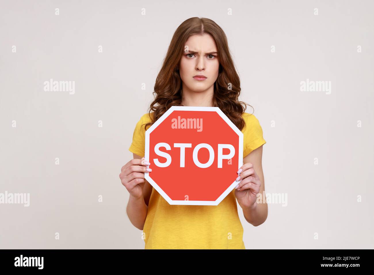 Interdiction, accès interdit. Jeune femme sérieuse en T-shirt jaune tenant un panneau d'arrêt rouge, avertissement et regardant avec colère et stricte à la caméra. Prise de vue en studio isolée sur fond gris. Banque D'Images