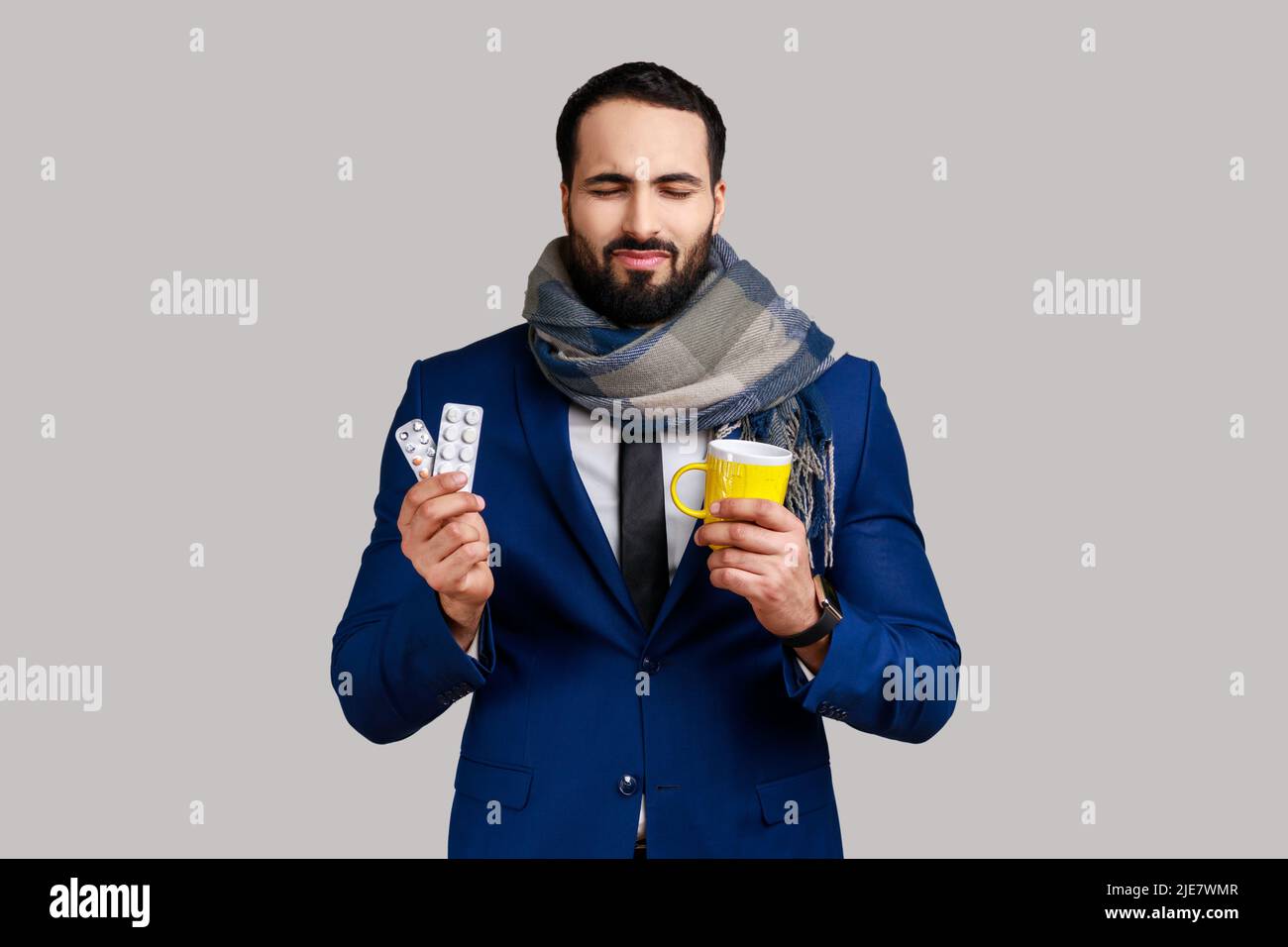 Homme barbu malsain debout et tenant une tasse jaune avec du tissu blanc, boire du thé chaud, a la grippe ou la grippe, portant le costume de style officiel. Prise de vue en studio isolée sur fond gris. Banque D'Images