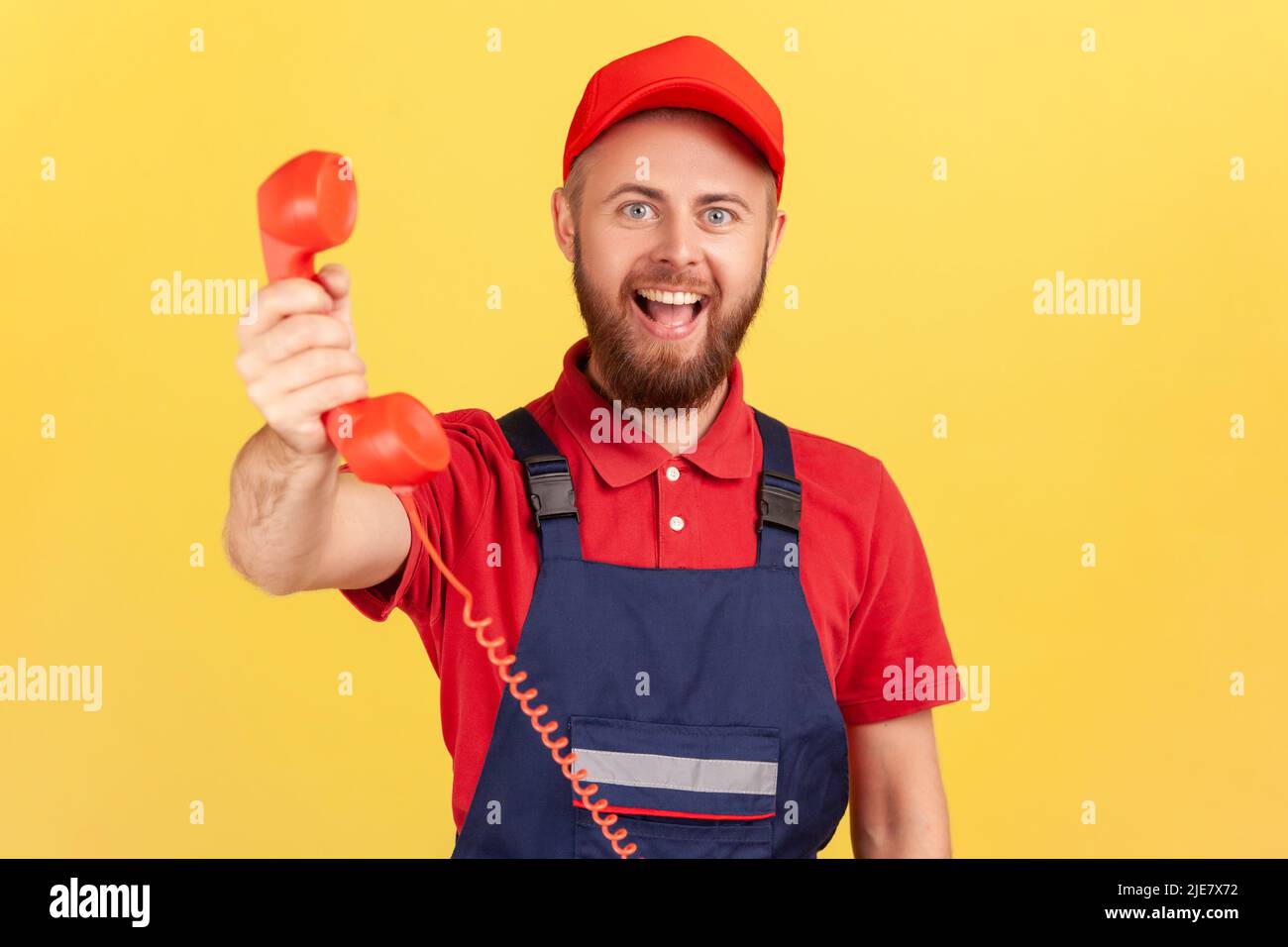 Portrait d'un homme à main positif portant un uniforme bleu tenant le combiné, lui demandant de l'appeler et de commander le service, regardant l'expression heureuse de l'appareil photo. Studio d'intérieur isolé sur fond jaune. Banque D'Images