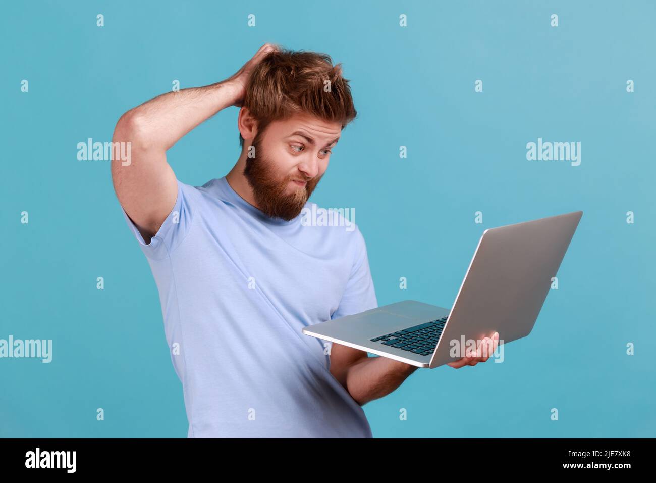 Portrait d'un homme barbu bien pensé debout et dactylographiant sur un ordinateur portable, ayant une nouvelle idée et planifiant sa propre stratégie, tenant une main sur la tête. Studio d'intérieur isolé sur fond bleu. Banque D'Images