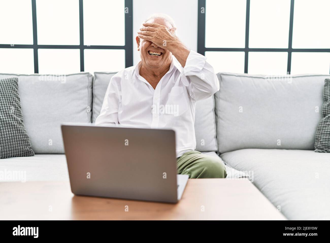 Homme senior utilisant un ordinateur portable à la maison assis sur le canapé en souriant et en riant avec la main sur le visage couvrant les yeux pour la surprise. Concept aveugle. Banque D'Images