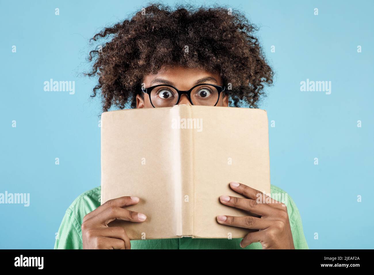 Portrait de l'homme américain africain émotionnel en lunettes couvrant le visage avec le livre, se cachant derrière lui avec les yeux grands ouverts Banque D'Images