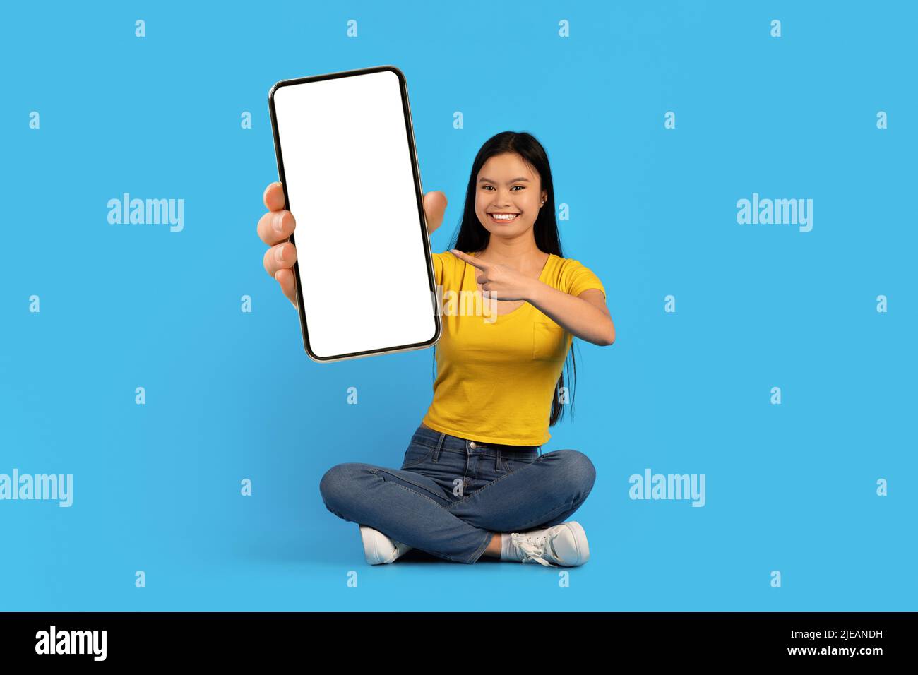 Une jeune femme chinoise souriante avec le syndrome de Down s'assoit sur le sol, pointant le doigt vers le téléphone avec un écran vierge Banque D'Images