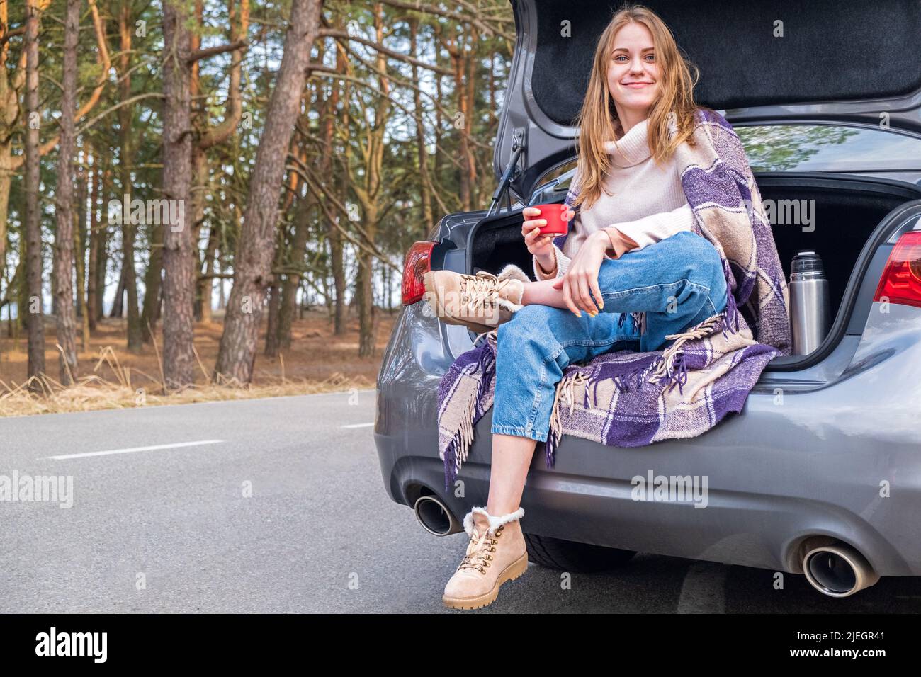 Une jeune fille imbéciforme et souriante regarde dans l'appareil photo assis dans le coffre d'une voiture avec une tasse de thé sur fond de printemps ou d'automne Banque D'Images