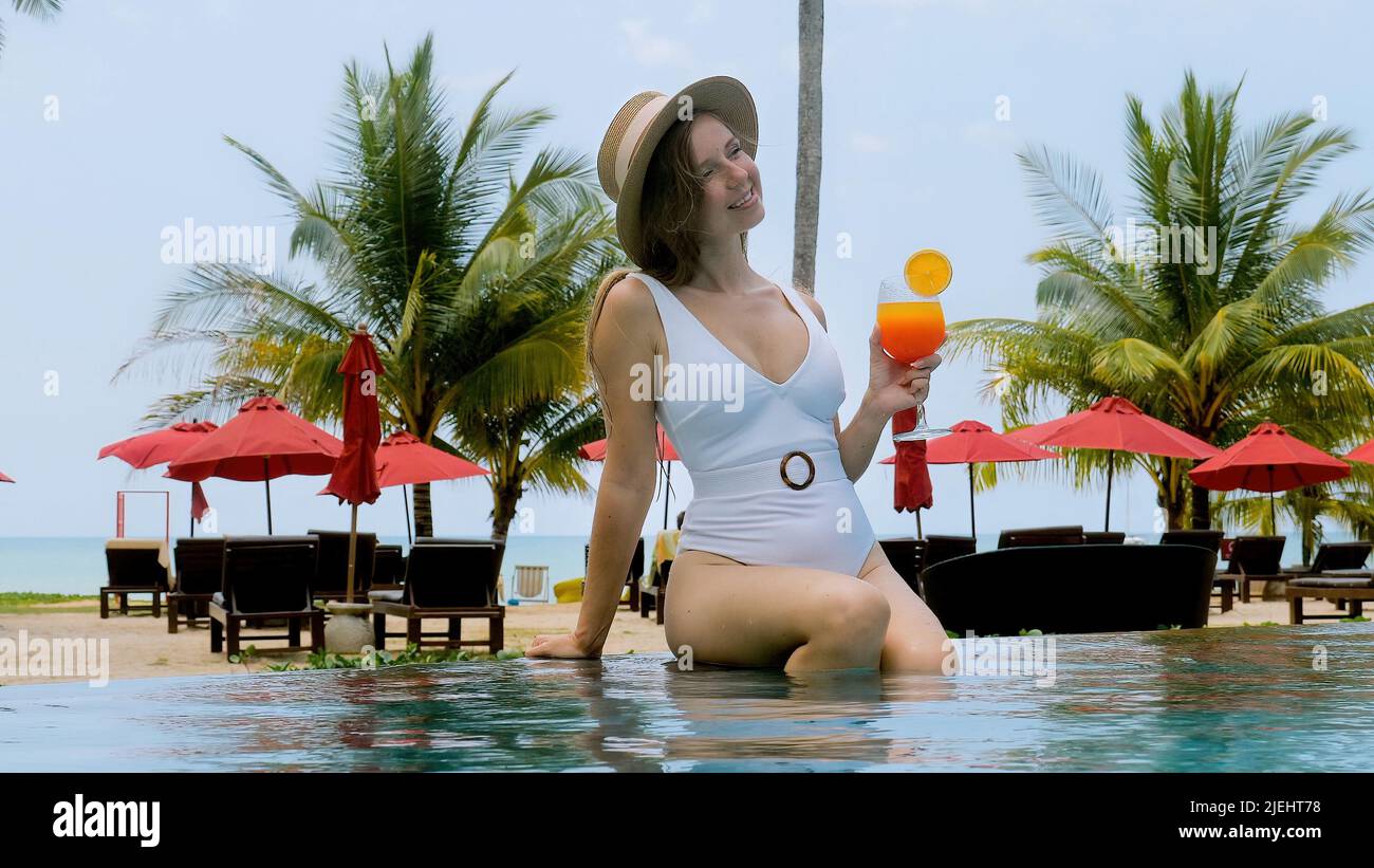 Belle fille sexy en maillot de bain blanc sourires Profitez de la détente avec un cocktail d'orange dans le Resort. Une jeune femme en chapeau de paille est assise sur le bord de l'extérieur Banque D'Images