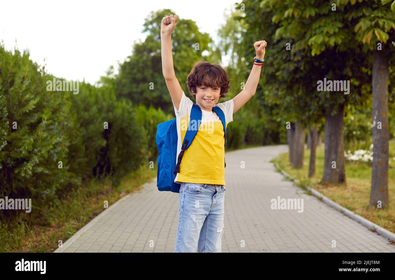 Joyeux garçon d'école gai debout dans le parc, levant les mains et souriant Banque D'Images