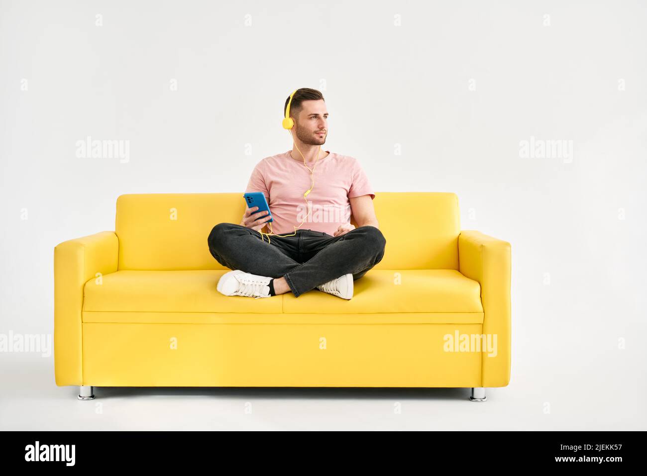 Décontracté jeune homme porter un casque écouter de la musique par son téléphone portable tout en étant assis sur un canapé jaune sur fond blanc Banque D'Images