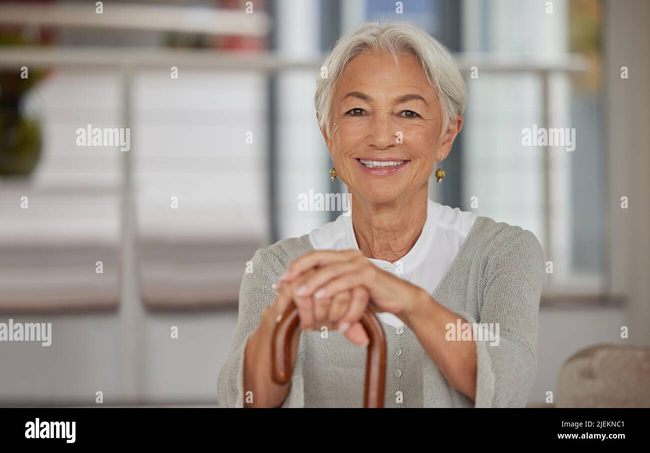 Portrait d'une femme âgée à la retraite tenant une canne en bois et souriant assis à la maison. Bonne vieille dame penchée sur un bâton de marche pour le soutien et Banque D'Images