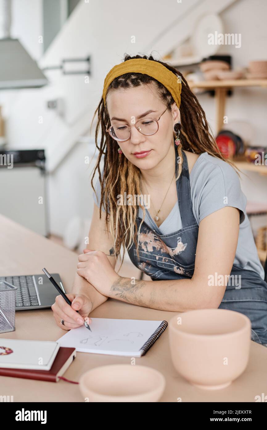 Jeune femme sérieuse se concentrant sur la création d'un nouveau dessin de faïence dans le bloc-notes tout en étant assise sur le lieu de travail en studio Banque D'Images