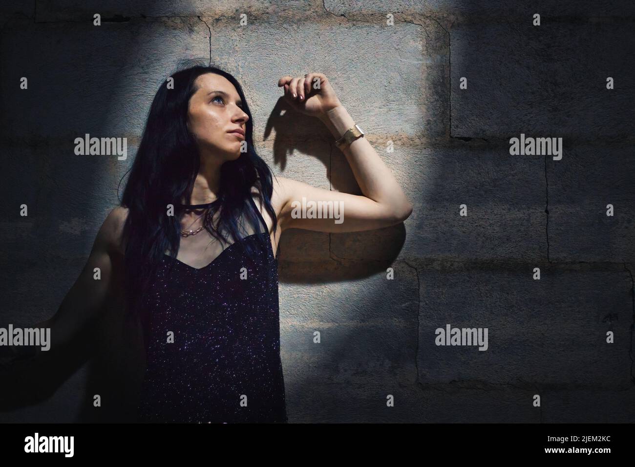 Une jeune femme attrayante portant une robe de soirée noire étincelante debout contre un mur regardant vers le haut dans un spot lumineux Banque D'Images