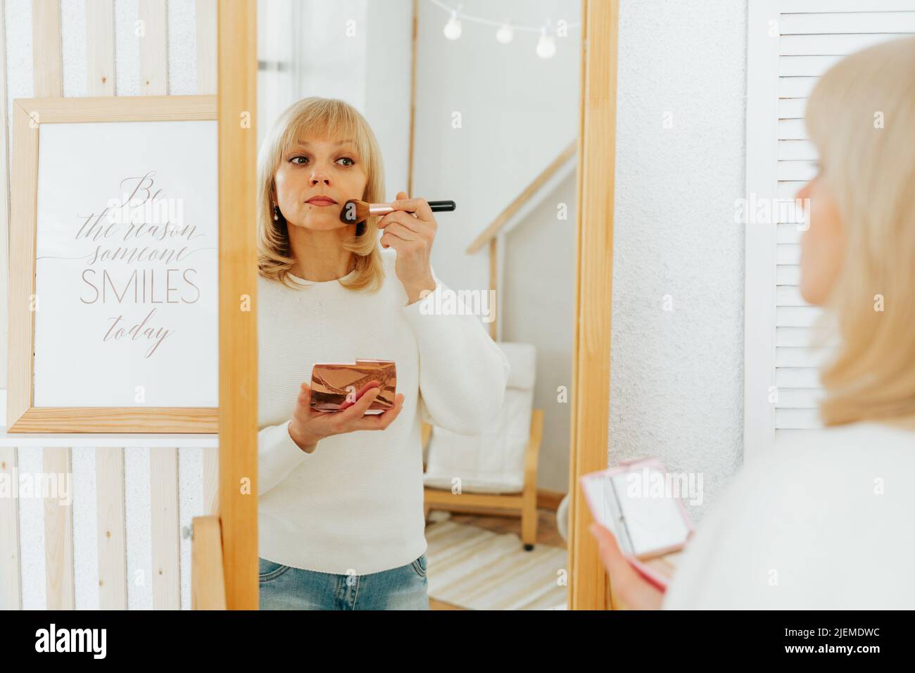 Belle femme caucasienne mûre et souriante avec des cheveux blonds debout près du miroir avec la palette de maquillage et la brosse, faisant le maquillage et regardant Banque D'Images