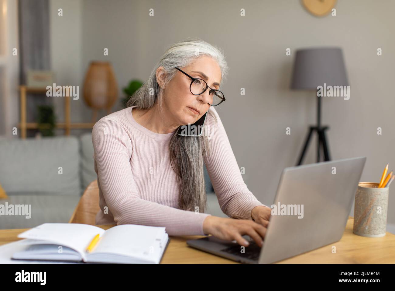 Une vieille dame européenne sérieuse avec des cheveux gris dans les lunettes travaille sur l'ordinateur et les appels par téléphone à l'intérieur de la chambre Banque D'Images