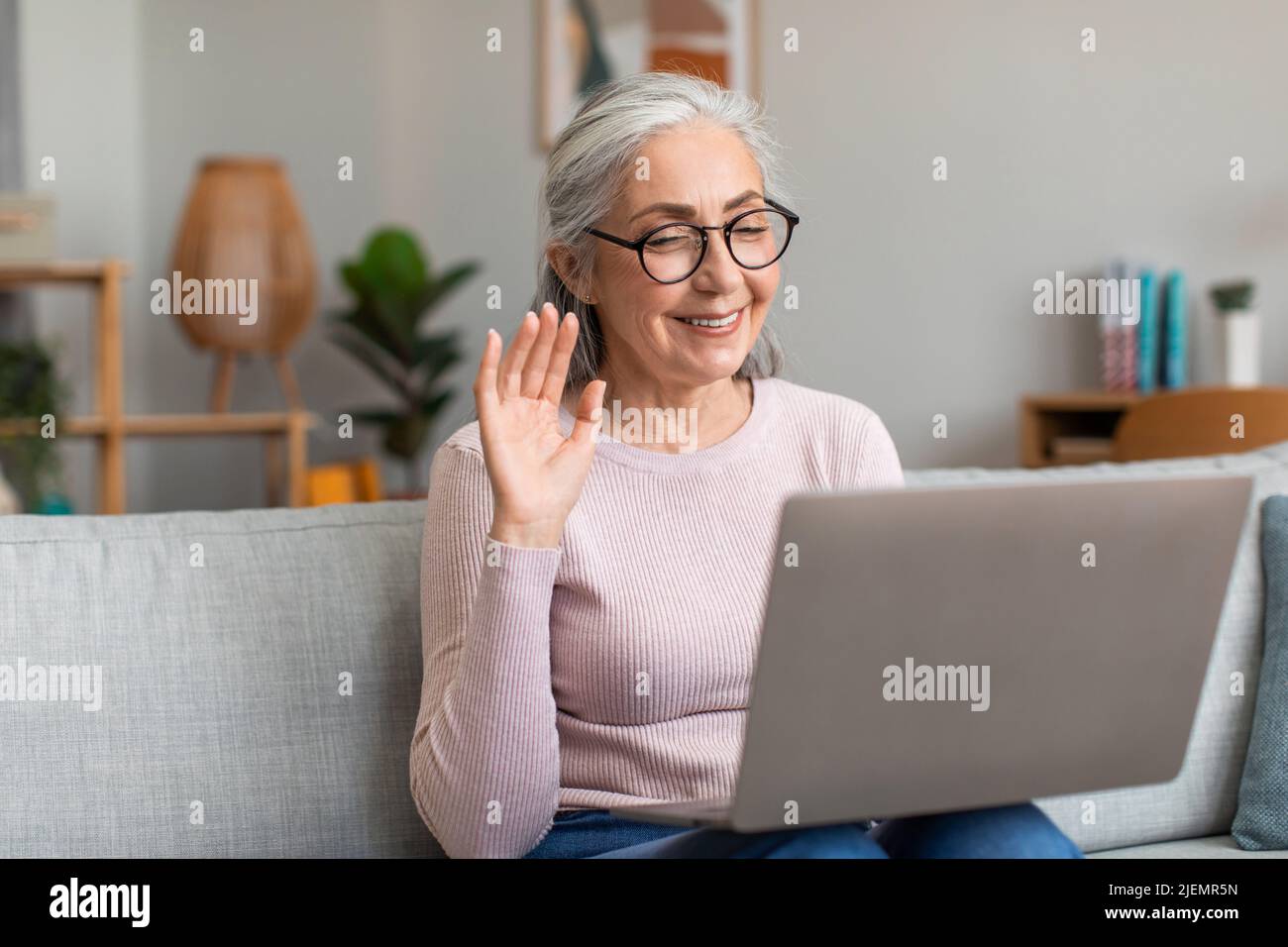 Contente européenne senior dame avec des cheveux gris dans les lunettes vagues sa main, regarde l'ordinateur dans l'intérieur de la salle de séjour Banque D'Images