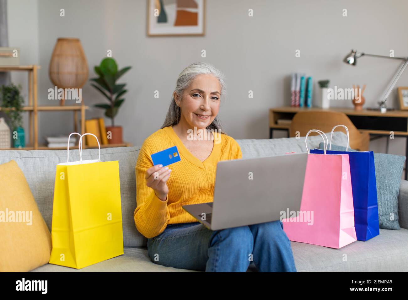 Femme senior européenne souriante avec cheveux gris avec des paquets après le shopping utilise carte de crédit et ordinateur portable pour le shopping en ligne Banque D'Images