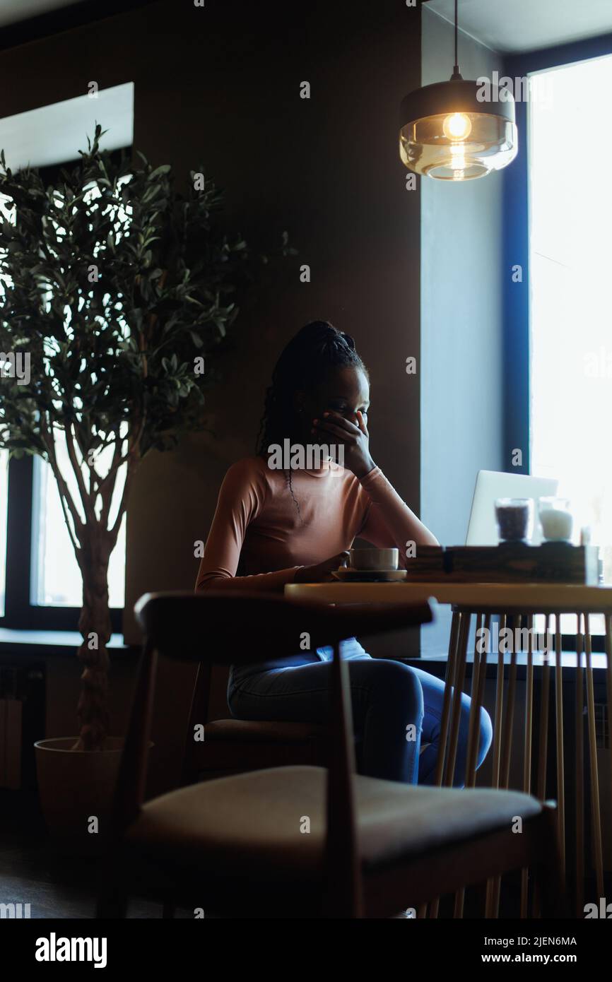 Portrait d'une jeune femme afro-américaine en train de rire assise à une table, regardant un ordinateur portable à écran, couvrant la bouche avec la main. Banque D'Images