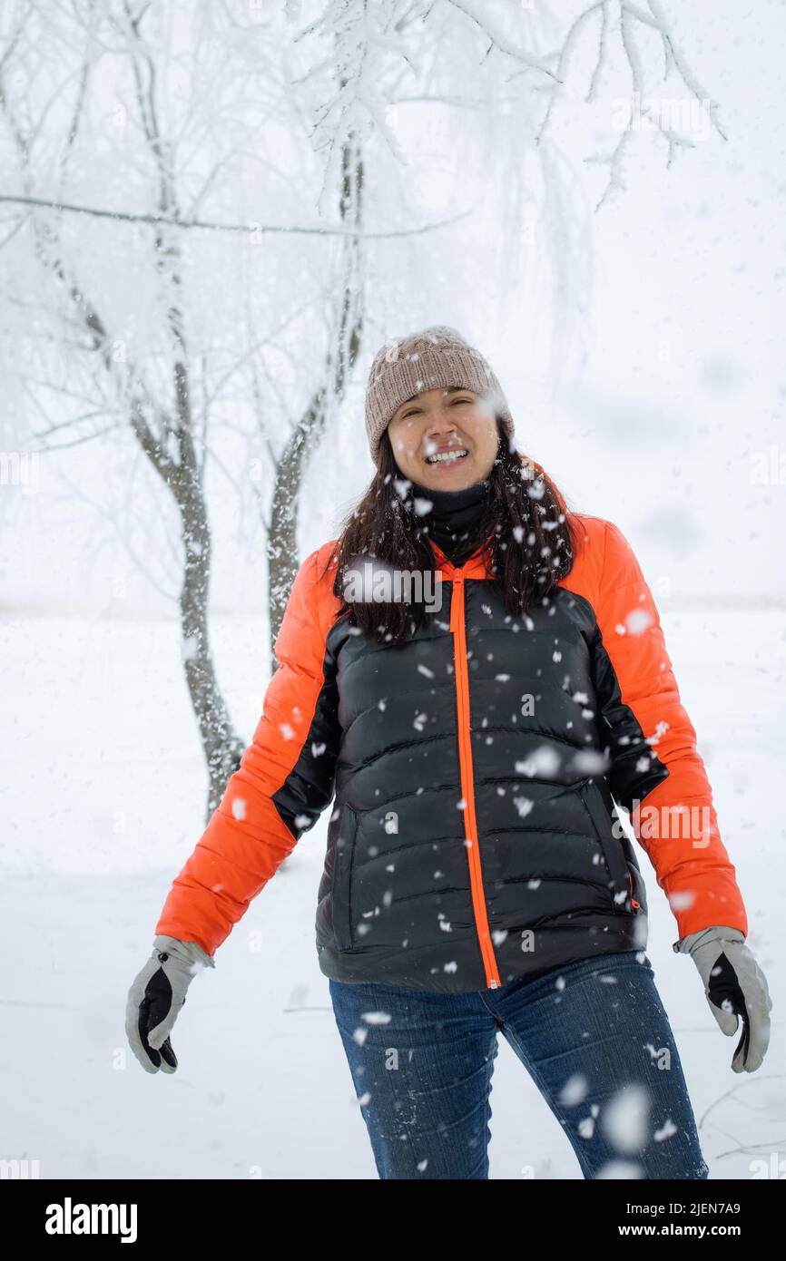 Portrait d'une femme d'âge moyen souriante vêtue chaleureusement debout sur un sol enneigé, regardant l'appareil photo avec les cheveux et le chapeau couverts de neige. Bel hiver Banque D'Images
