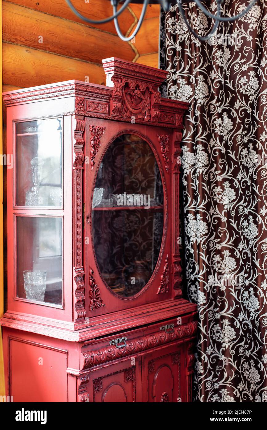 Armoire vintage en bois rouge avec ornements sculptés dans le coin de la maison rurale en bois entre les fenêtres près du rideau. Banque D'Images