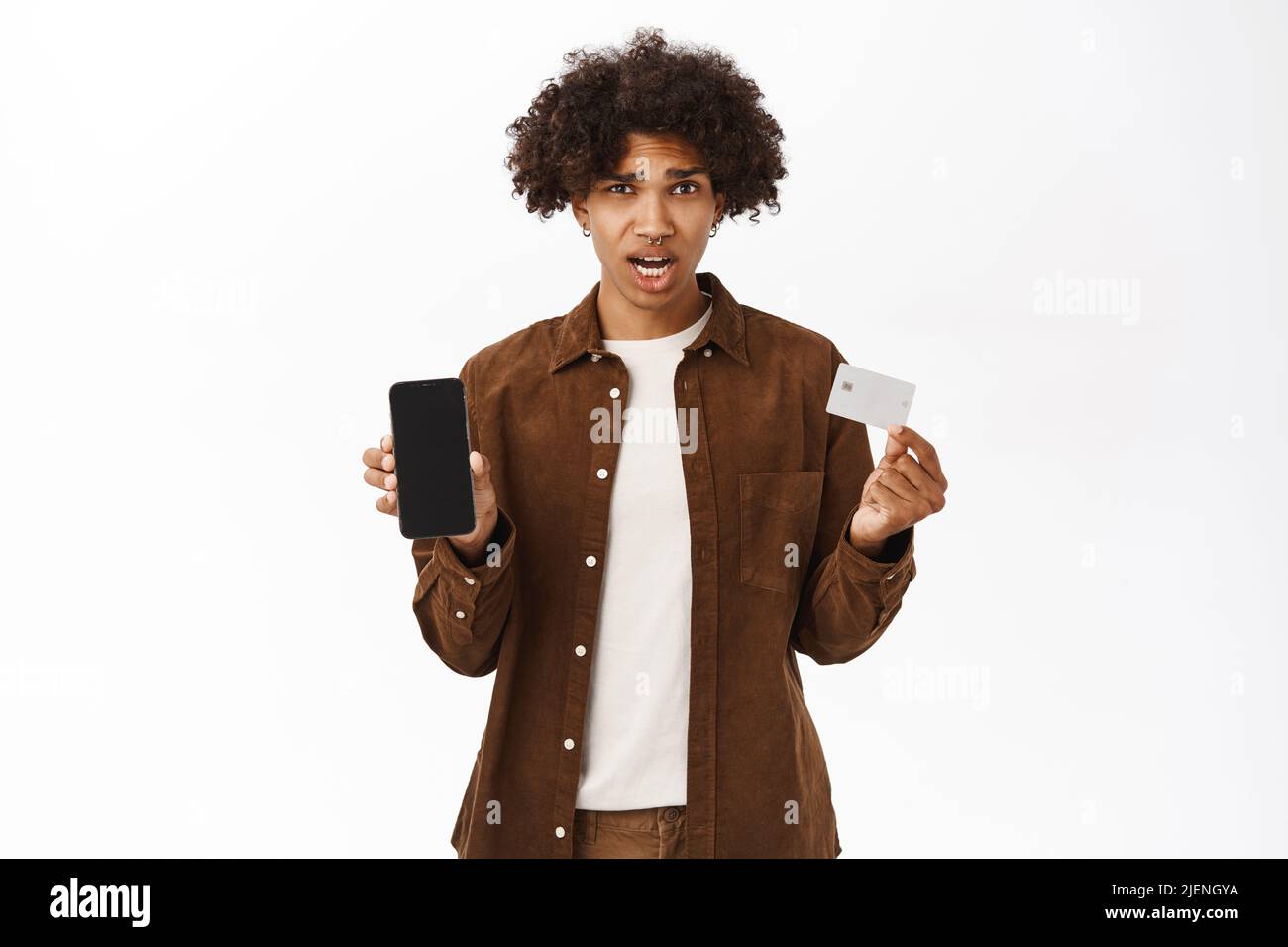 Portrait d'un homme hispanique choqué montre carte de crédit et téléphone mobile, application écran, debout dans des vêtements décontractés sur fond blanc Banque D'Images
