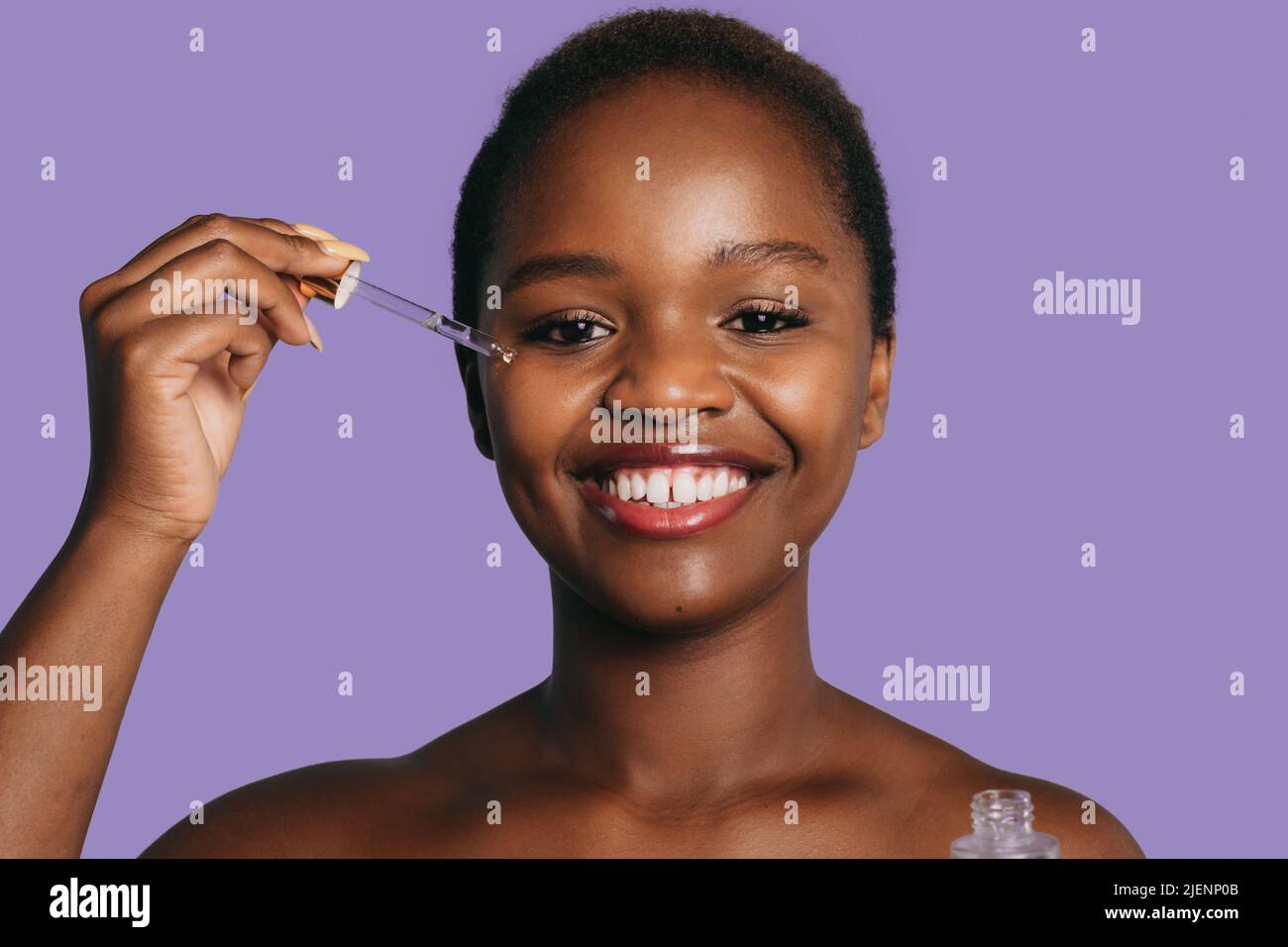 Femme afro souriante appliquant du sérum hydratant sur sa peau isolée sur fond violet. Belle femme noire. Beauté naturelle. Soin du visage Banque D'Images