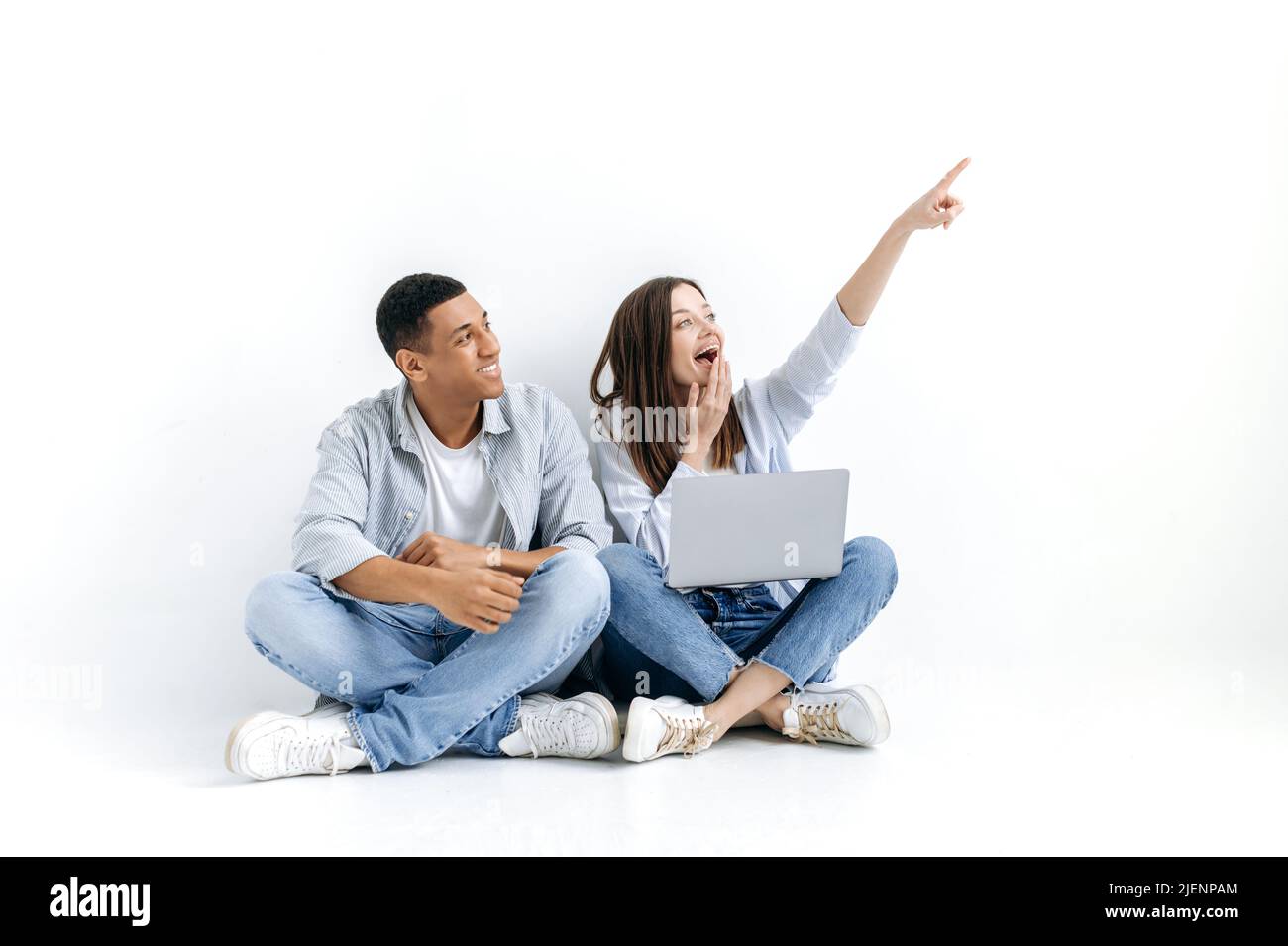 Un couple multiracial positif, une femme caucasienne et un homme hispanique, sont assis avec un ordinateur portable sur le sol sur fond blanc isolé, regardant sur le côté, la fille pointe avec sa main à quelque chose Banque D'Images