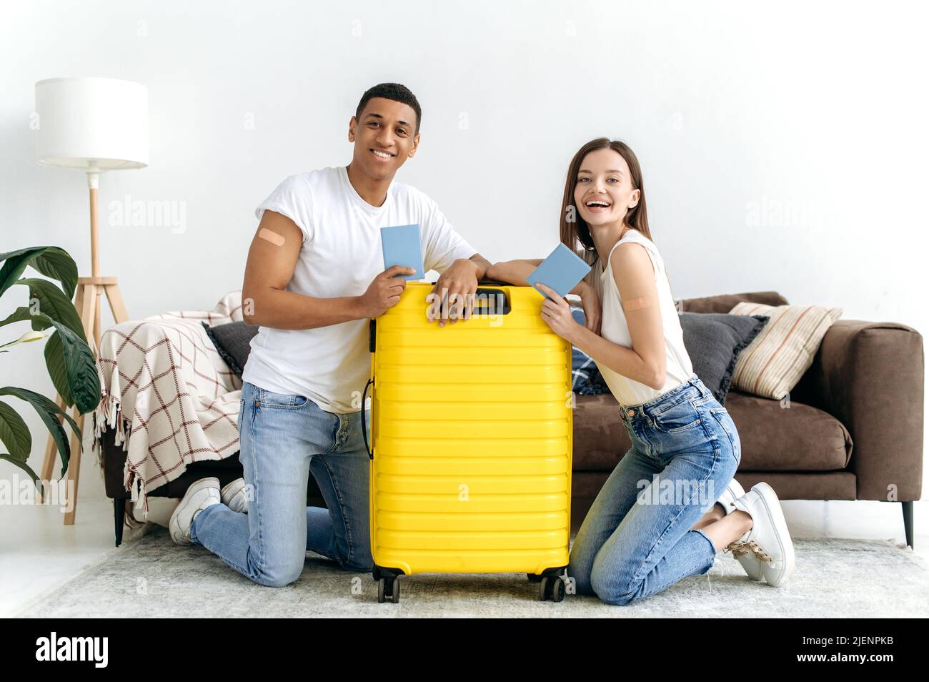 Vacances, voyage longtemps attendus. Heureux couple multiracial, a reçu le vaccin et en voyage, s'asseoir sur le sol dans le salon près de la valise jaune, tenir leur passeport, regarder l'appareil photo, sourire Banque D'Images