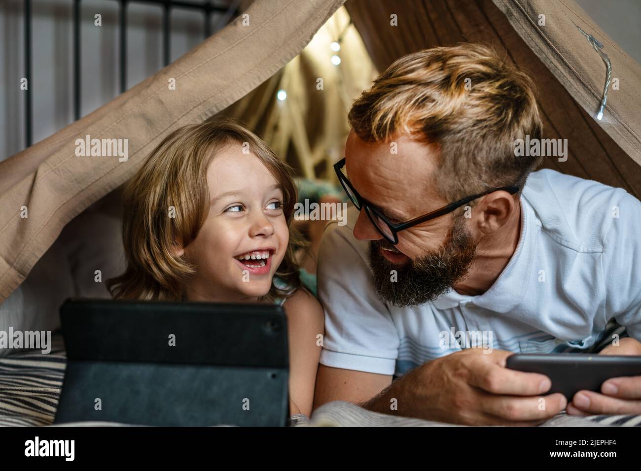 Portrait d'un garçon de 6 ans et de son père qui s'amusent à jouer dans une tente à tipi. Père et fils utilisant une tablette numérique pour regarder des dessins animés ou jouer à des jeux informatiques dans une tente pour enfants à la maison. Banque D'Images