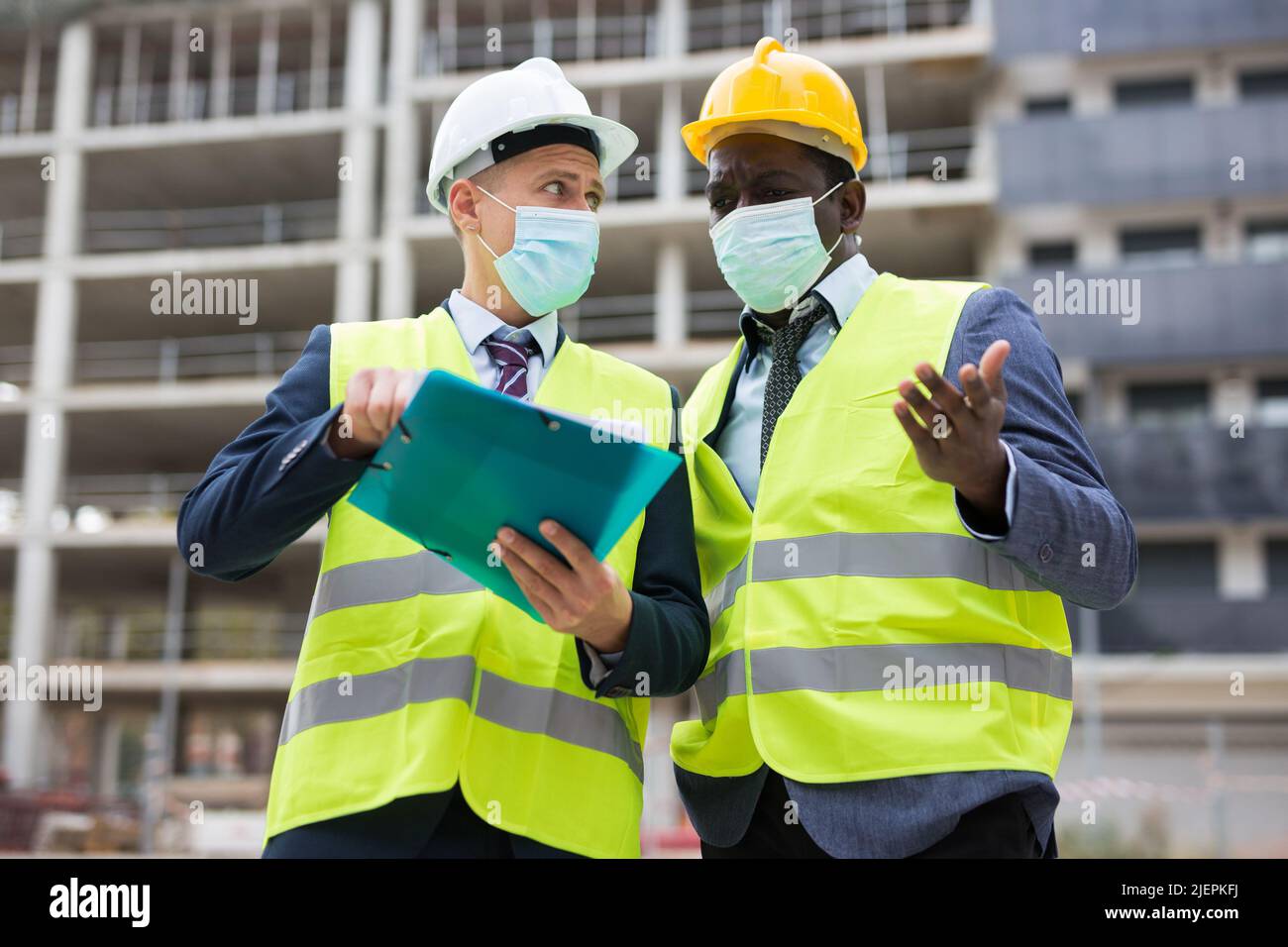 Deux ingénieurs en masques de protection, travaillant sur un chantier de construction pendant une pandémie, discutent d'un plan de construction, en tenant un ..estimate entre leurs mains Banque D'Images