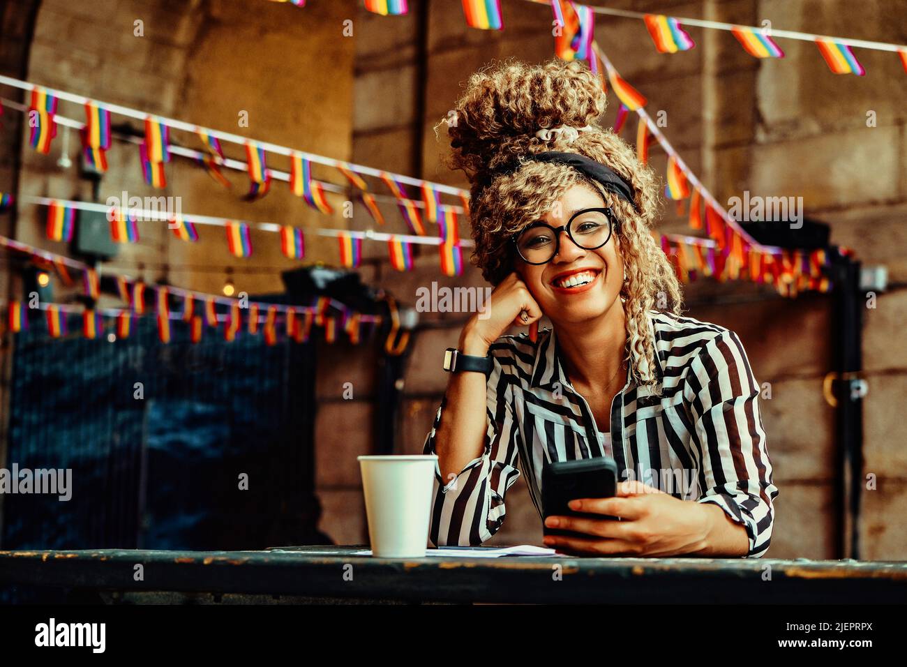 Portrait d'une femme souriante avec une coiffure afro, des lunettes, un smartphone et une tasse de papier regardant l'appareil photo assis dans un café Banque D'Images