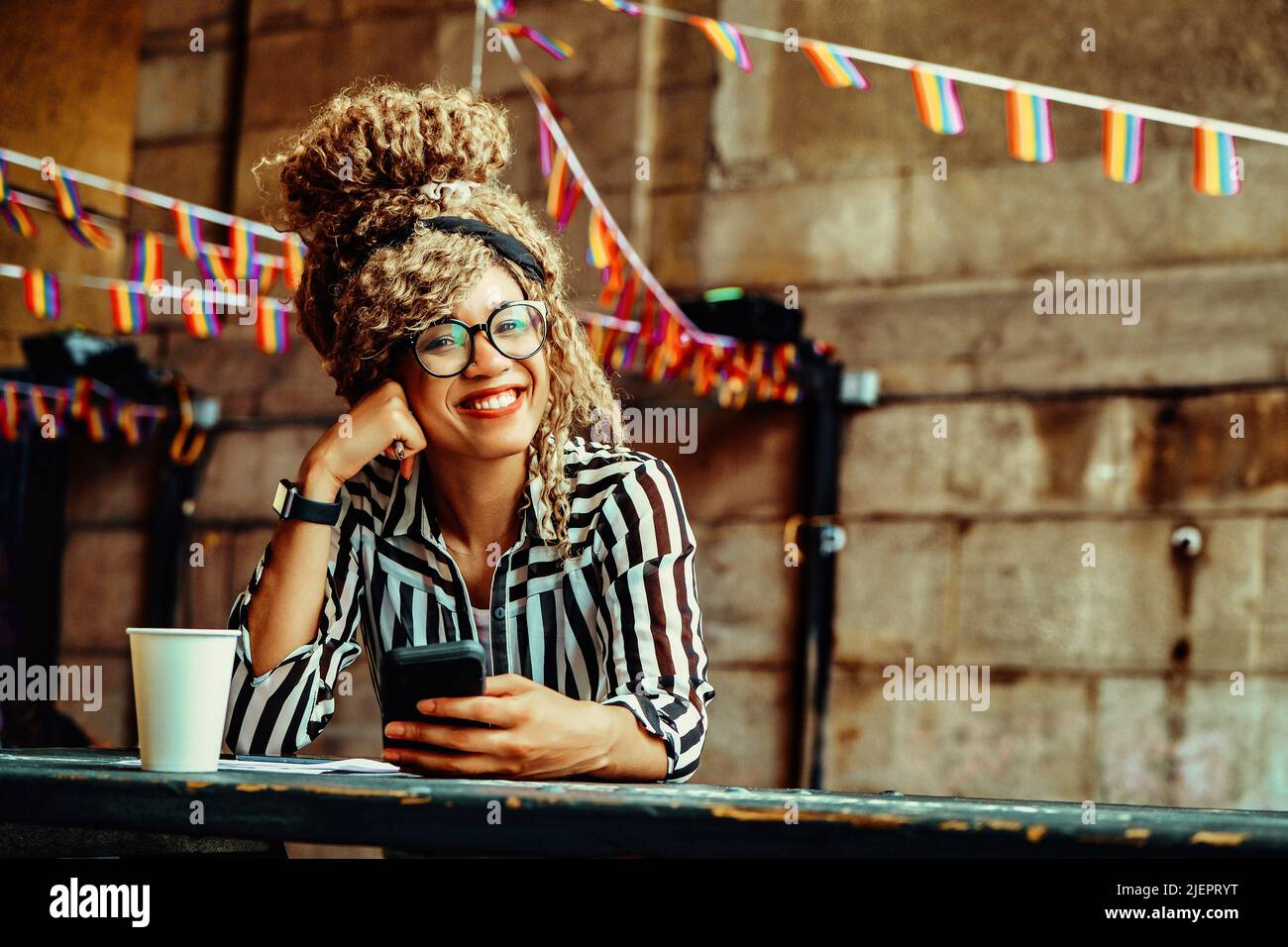 Portrait d'une femme souriante avec une coiffure afro, des lunettes, un smartphone et une tasse de papier regardant l'appareil photo assis dans un café Banque D'Images