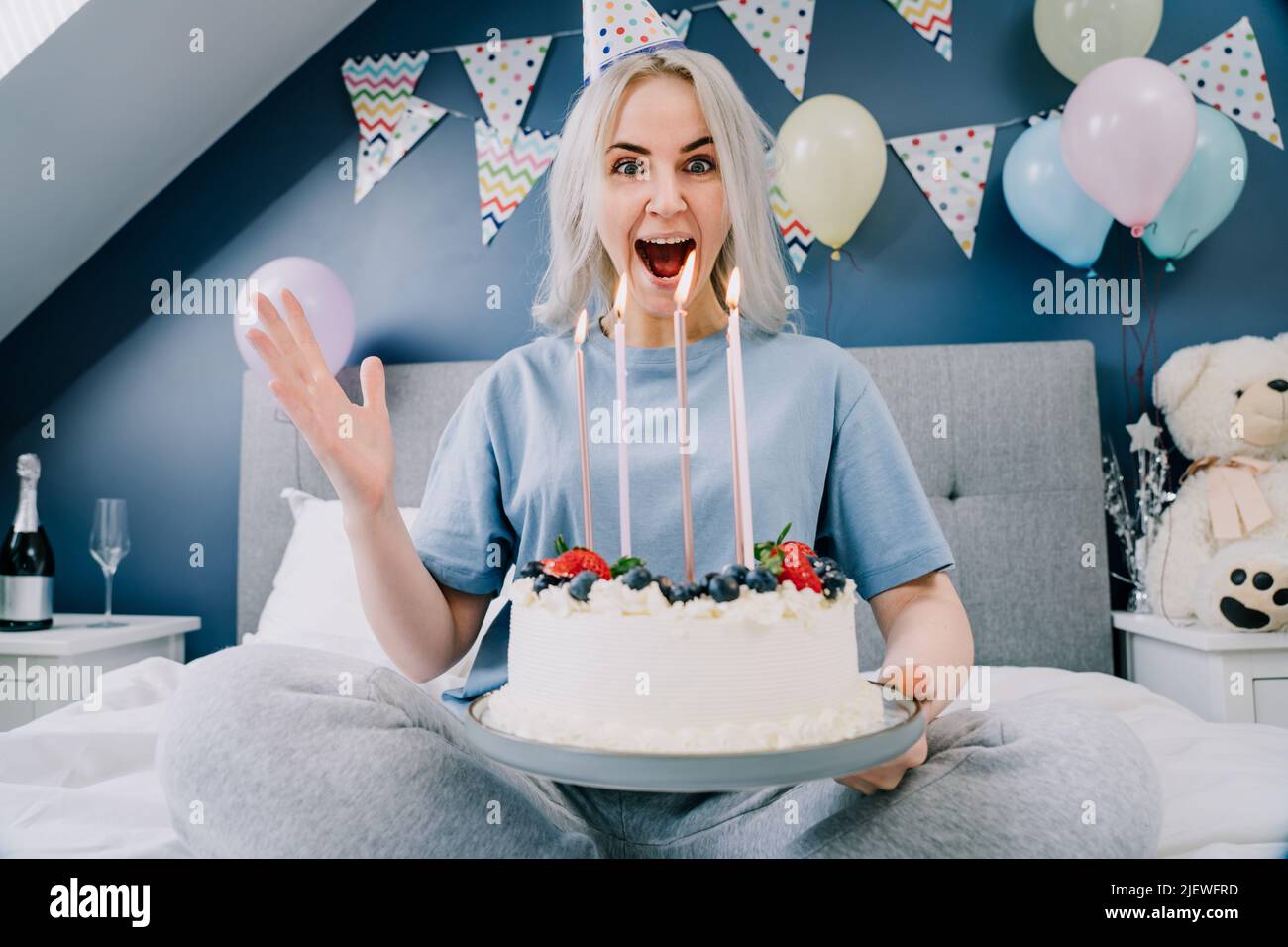 Femme émotive et excitée dans un chapeau d'anniversaire tenant un gâteau de fête avec des bougies, tout en étant assise sur le lit dans la chambre décorée. Surprise du matin. Bonne birt Banque D'Images Femme émotive et excitée dans un chapeau d'anniversaire tenant un gâteau de fête avec des bougies, tout en étant assise sur le lit dans la chambre décorée. Surprise du matin. Bonne birt Banque D'Images