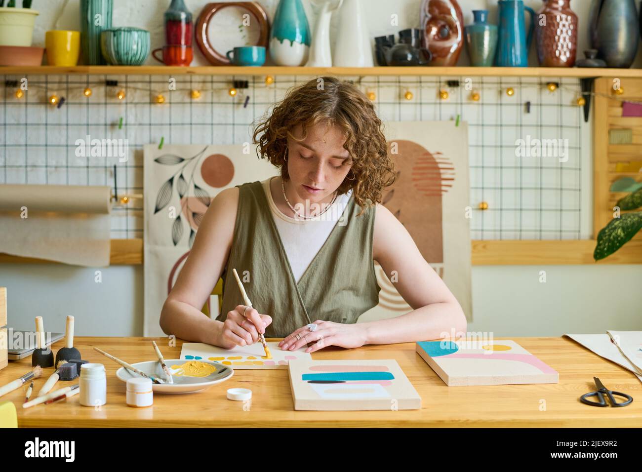 Jeune femme créative en vêtements décontractés avec pinceau et gouache pour créer une nouvelle peinture tout en étant assise sur le lieu de travail en studio Banque D'Images