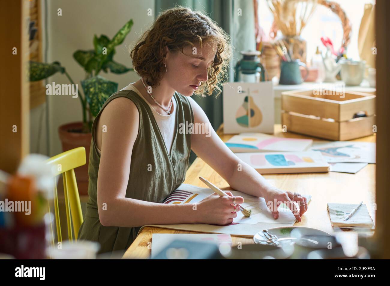 Jeune femme sérieuse avec pinceau assis au travail en studio et peindre des choses abstraites avec des aquarelles ou gouache sur papier Banque D'Images
