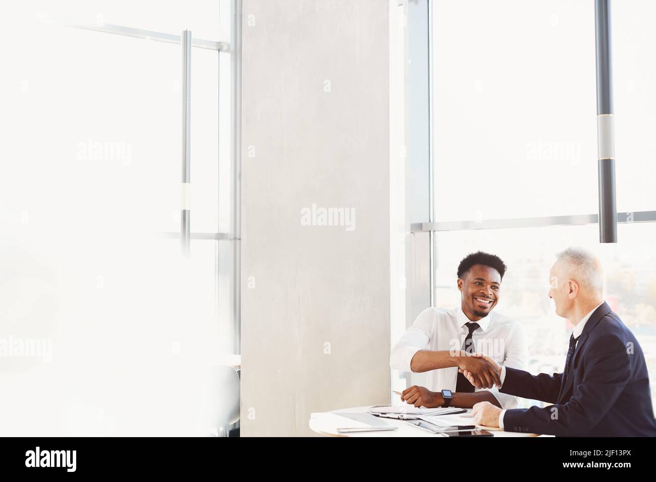 Des hommes d'affaires multiethniques gaies en costume formel assis à table et faisant poignée de main tout en atteignant un accord Banque D'Images