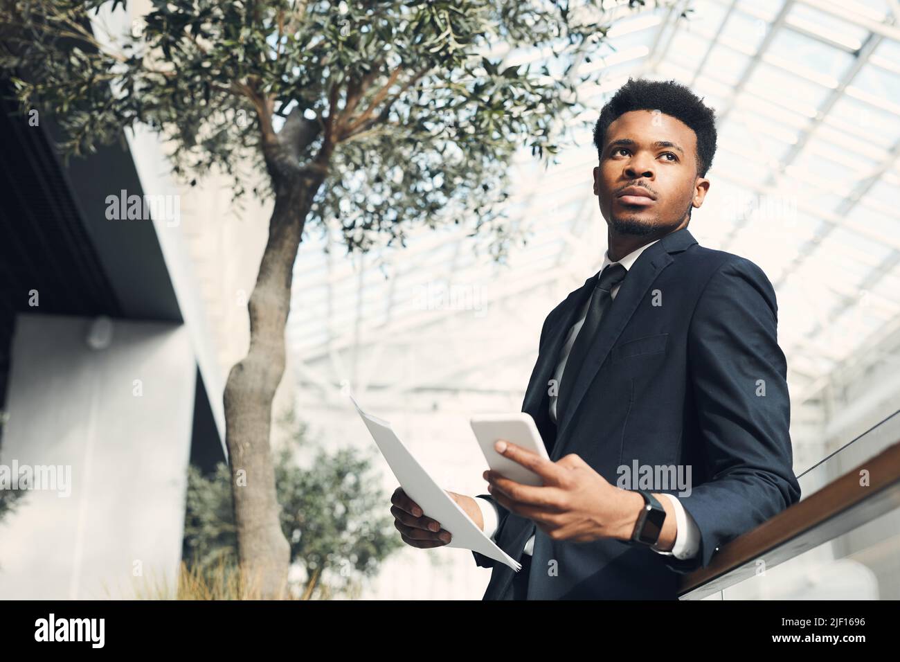 Un jeune homme d'affaires noir plein de confiance dans une veste habillée penchée sur une rampe dans un hall moderne avec des arbres et planifiant un projet commercial rentable Banque D'Images