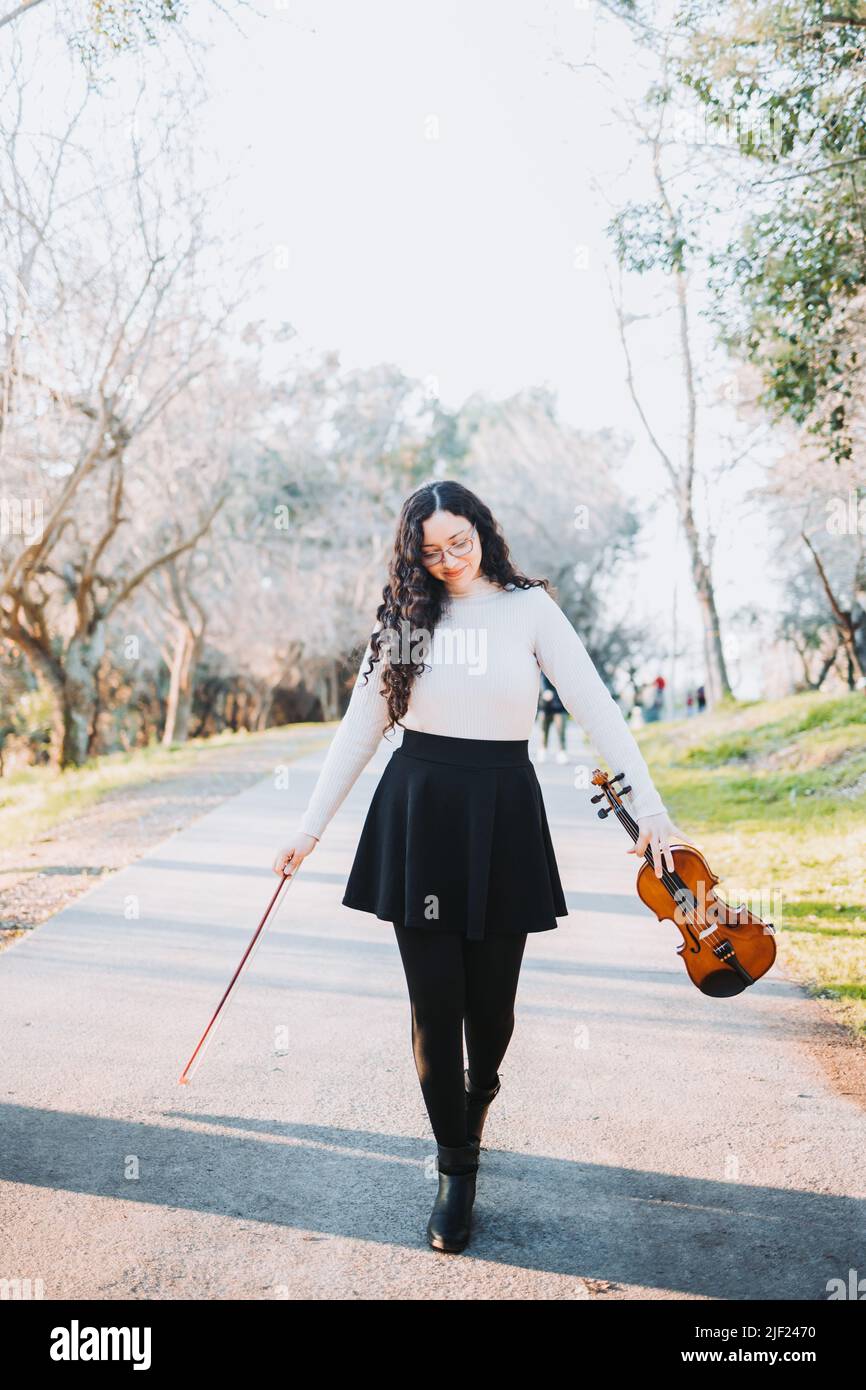 Belle femme brune tenant un violon et un arc, marchant dans la route du parc au coucher du soleil. Banque D'Images