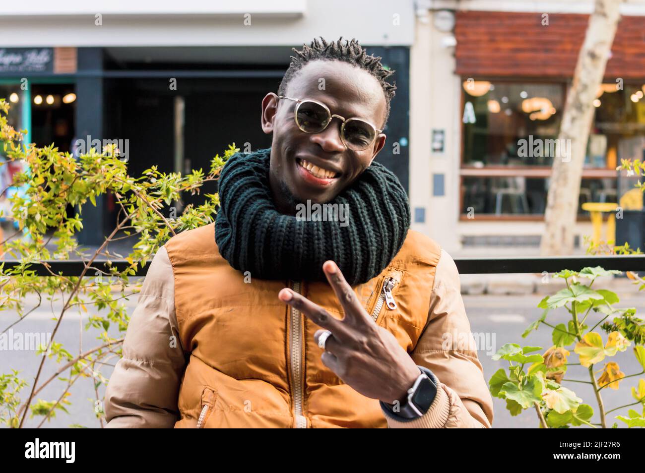 portrait jeune africain portant des lunettes et des vêtements d'hiver, assis à l'extérieur du restaurant faisant le signe v avec les mains regardant l'appareil photo et souriant, fron Banque D'Images