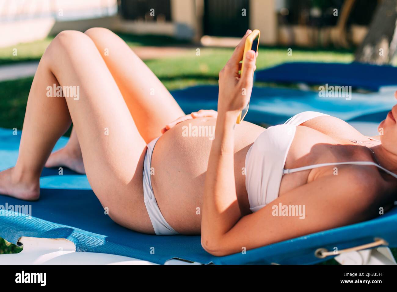 jeune femme enceinte allongé sur un transat au bord de la piscine, bain de soleil tout en discutant sur son smartphone Banque D'Images