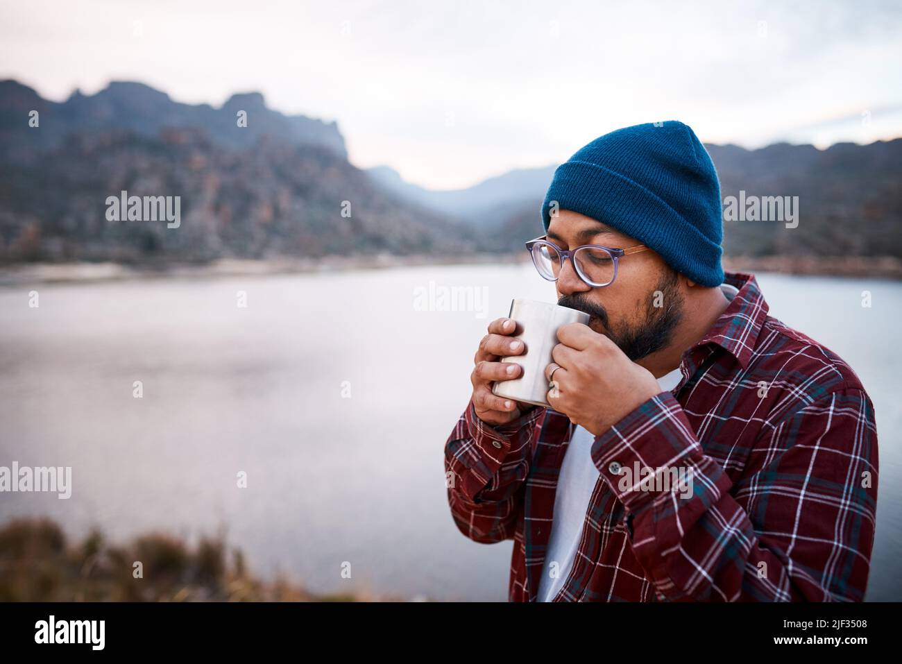 Un homme d'Asie du Sud soupière le café d'une tasse de camping avec vue sur les montagnes et le lac Banque D'Images Un homme d'Asie du Sud soupière le café d'une tasse de camping avec vue sur les montagnes et le lac Banque D'Images