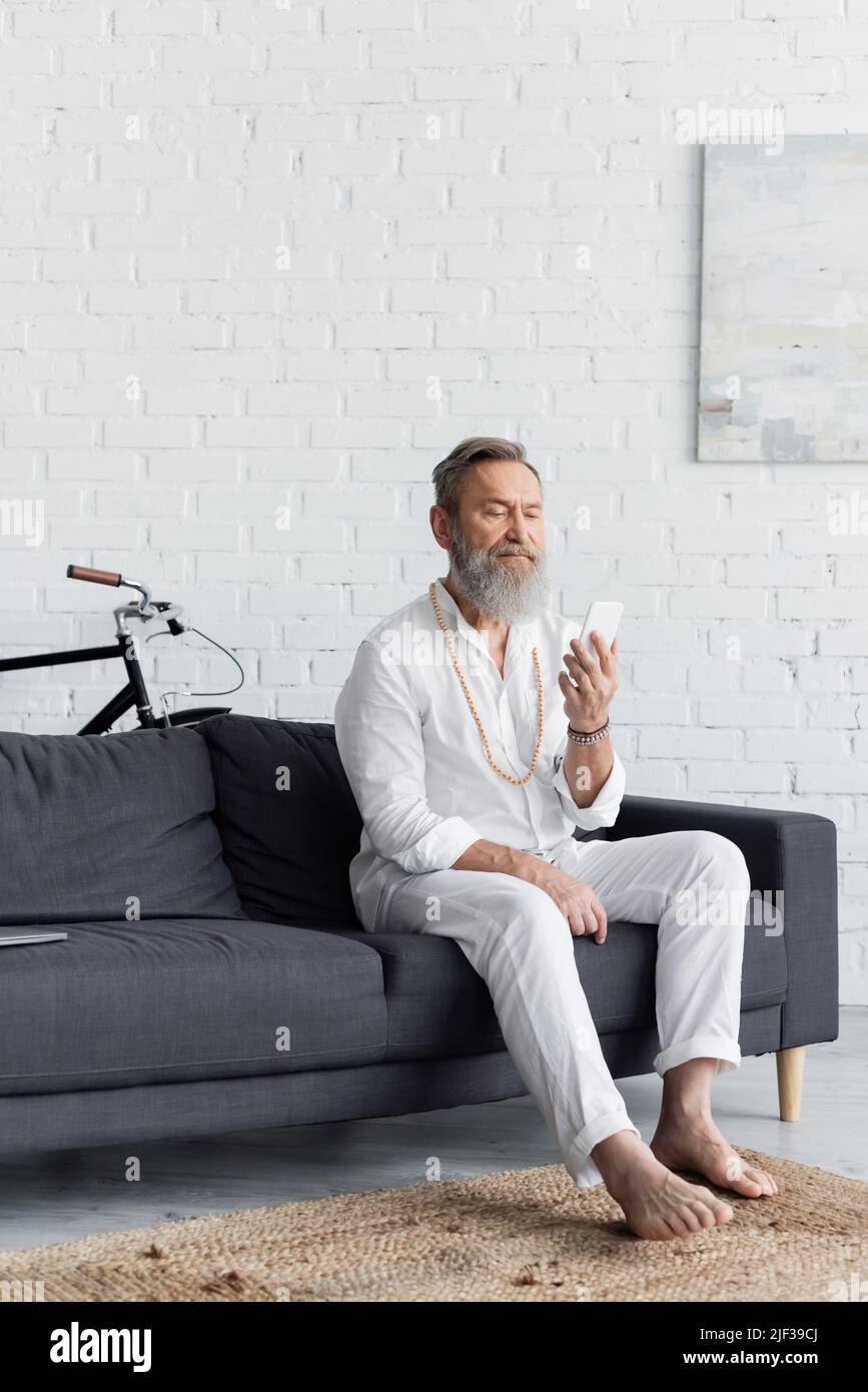 homme expérimenté gourou pieds nus avec téléphone mobile assis sur un canapé à la maison Banque D'Images