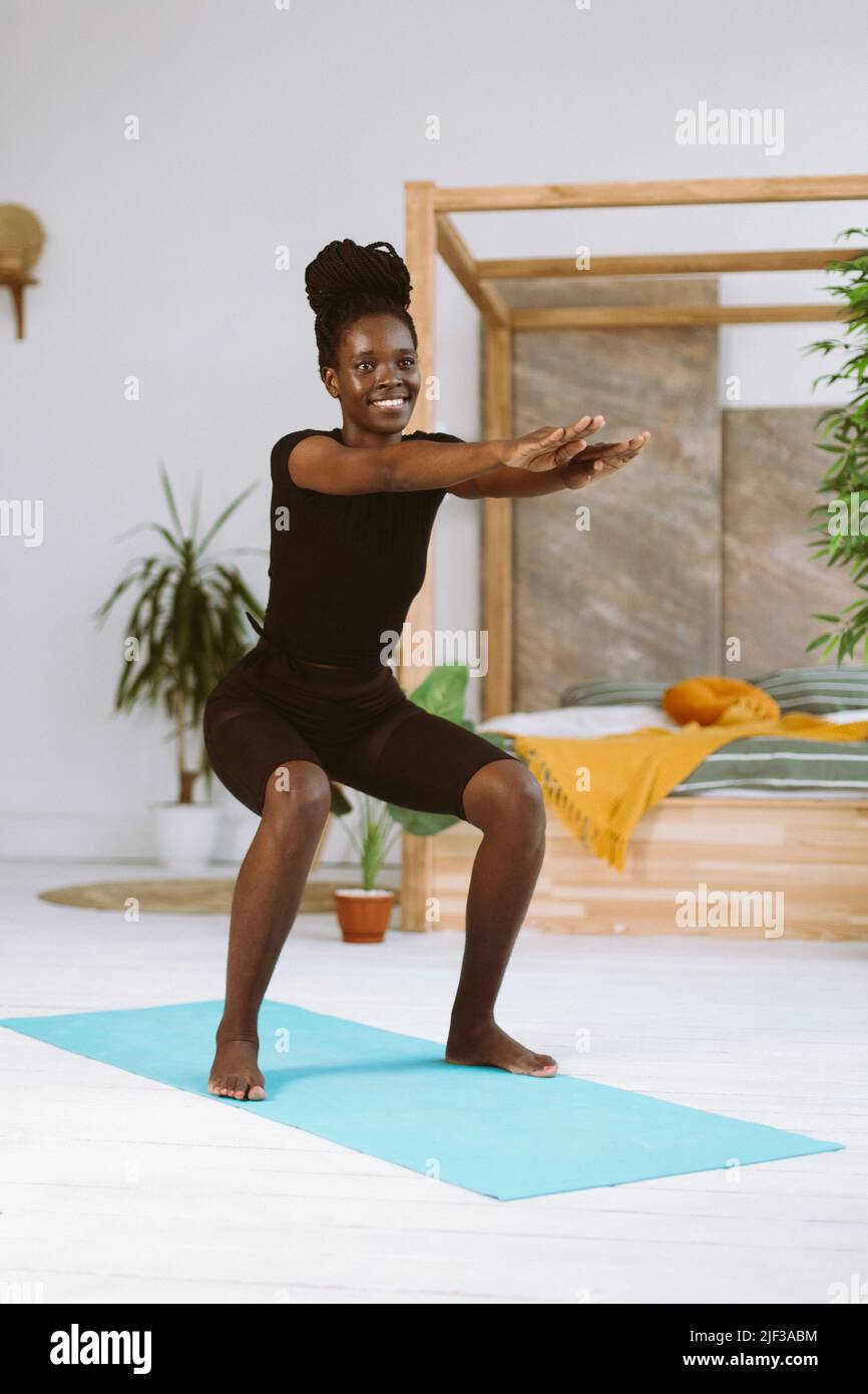 Athlétique afro-américaine femme avec sourire en squatant avec les bras étendus vers l'avant, entraînement sur le tapis de gymnastique dans un studio décoré. Pompage musculaire, sport Banque D'Images