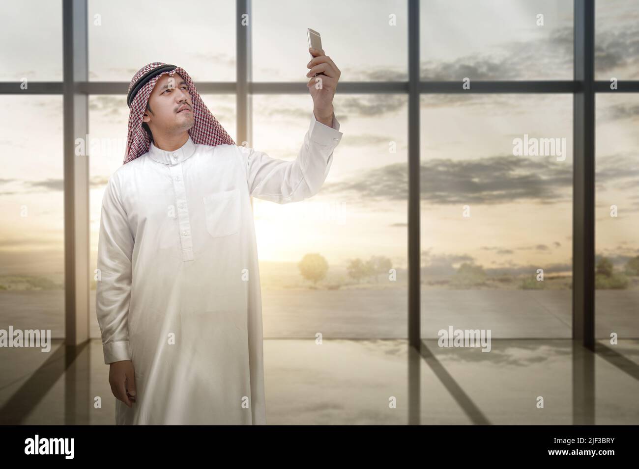 Homme arabe portant le keffiyeh regardant le téléphone mobile à l'intérieur de la pièce avec verre de fenêtre et coucher de soleil ciel fond Banque D'Images